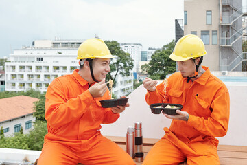 Smiling builders sitting on house rooftop outdoors and eating noodles for lunch