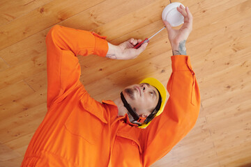 Technician with screwdriver installing smoke detector on wooden ceiling