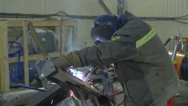 A welder in protective clothing and a mask works with a welding machine in a large cold garage