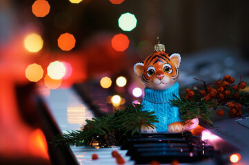 Obraz premium Christmas tree toy of a handsome tiger in a sweater on the piano keys. Colored lights of an electric garland. Branches of a Christmas tree and rowan berries. Happy New Year. Selective focus.