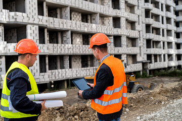 young strong hardy construction workers in overalls standing near a new building with a plan