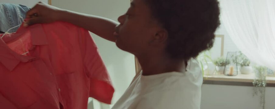 Young cheerful African American woman looking in mirror, smiling and choosing outfit at home