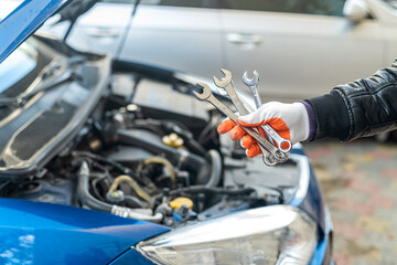 Man's gloved mechanic's hand holds car tools near a painted car with an open hood
