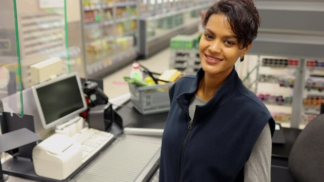 Young woman cashier posing for camera and smiling while sitting at checkout of store spbi. Closeup view of beautiful American female worker looks ahead with smile and poses, sits at workplace with