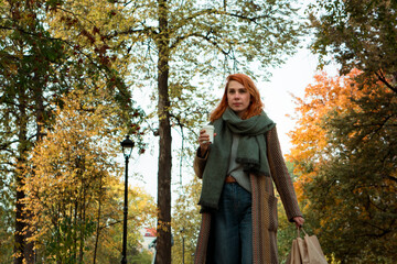 Fototapeta na wymiar Redhead young woman in stylish autumn outfit with paper eco shopping bags and cup of coffee walking in city park in fall