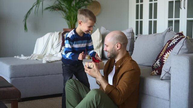 Fathers day celebrating at home. Adorable toddler boy presents gift and plays with loving parent on floor in contemporary living room spbd