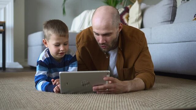 Father spends time with little son. Happy toddler boy and surprised spbd bald parent watch cartoons on tablet lying together on floor by sofa closeup