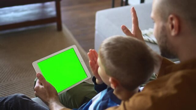 Father and son at videochat. Family of little boy and male parent sends air kisses at videocall via tablet with green screen on sofa at home closeup spbd