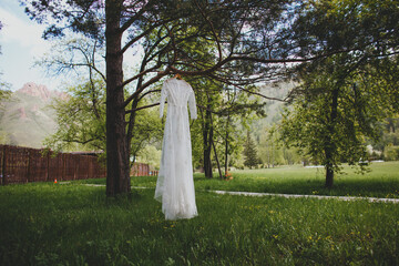 Wedding dress hanging on tree on background of wooden structure among summer greenery. High quality photo