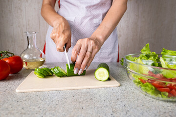 Female hands hold a knife and cut a cucumber for salad making. Cooking salad in the kitchen against the background of a woman in an apron.