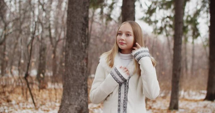Beautiful young woman in white coat touching hair in winter forest. Fashion model woman in winter coat posing in snowy forest