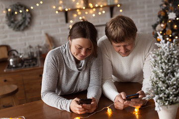 Couple in love (woman and man) using phones for texting or online shopping at home in Christmas decorated kitchen