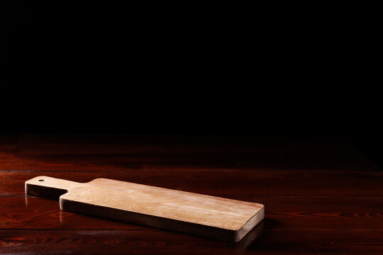 A clean empty light wooden cutting board lies on a dark wooden table. Black background. Angle view. Selective focusing.