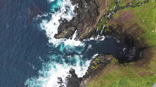 Aerial view of waves break on rocks of Faroe islands cliffs in a blue ocean.Drone Aerial Footage of green nature and the ocean.