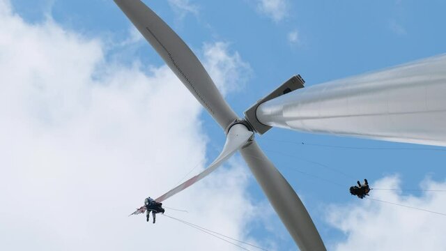 Professional high-altitude installers with climber equipment serve modern large wind generator blades under blue sky with white clouds low angle shot