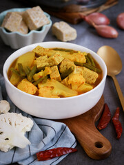 A bowl of Thai yellow curry mixed assorted vegetables such cauliflower, green bean, papaya with tempeh as a vegan dish. Ingredients such red dried chili and shallots. An alternate high protein dish