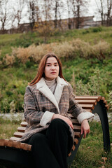 Pensive, thoughtful young woman in coat sitting alone on wooden bench. Sad woman daydreaming, thinking in autumn park. Autumn blues, feeling down. Waiting for someone. Urban casual outfit. Lifestyle