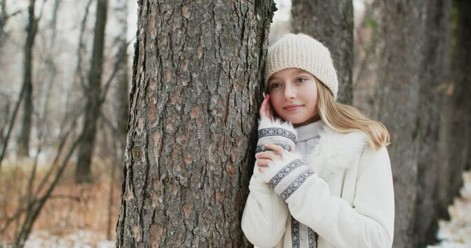 Romantic woman in white jacket and knitted hat leaned on tree trunk posing for shooting. Portrait stylish girl in winter clothes and hat posing in winter forest