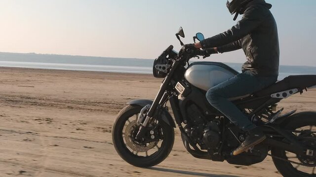 Motorcyclist driving his customized fast motorbike on the dirt road in desert around sea or lake. Slow motion