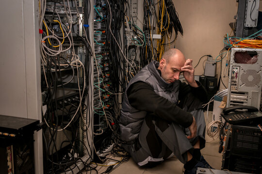 A frustrated technician sits in a server room. A man sits on the floor in a data center among many wires.