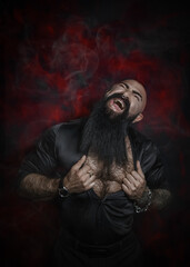 Stylish handsome man with a black beard in a black shirt with a naked torso and hairy chest screams against a red background of fire