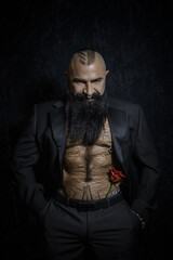 Stylish handsome man with a black beard in a black suit with a naked torso and hairy chest stands with a beautiful rose in his pants