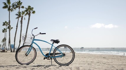 Blue bicycle, cruiser bike by sandy ocean beach, pacific coast, Oceanside pier California USA. Summertime vacations, sea shore. Vintage cycle, palms, sky, lifeguard tower watchtower hut, car truck. - obrazy, fototapety, plakaty
