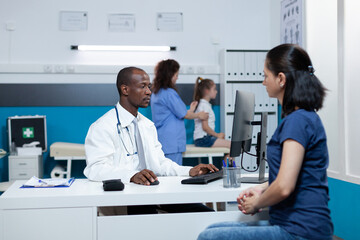 Fototapeta premium African american pediatrician doctor discussing illness expertise results with mother typing healthcare treatment on computer keyboard. Physician man with stethoscope working in hospital office
