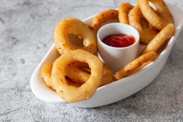 Battered onion rings in a dish,  with a tomato ketchup dip. On a concrete background