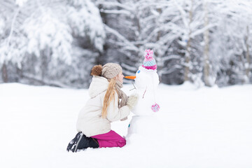 Blond caucasian girl wearing warm knitted clothes building a snowman on winter snowy day. Happy...