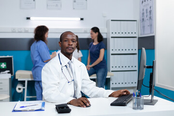 Portrait of african american pediatrician doctor sitting at desk table in hospital office analyzing disease expertise on computer during clinical examination. Pediatric man typing illness symptoms