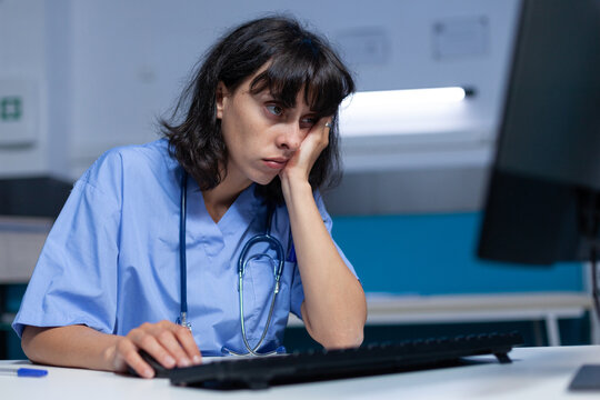 Medical specialist falling asleep while using computer, working late at night. Assistant feeling exhausted after doing overtime work on monitor for healthcare system. Sleepy nurse