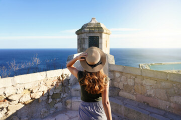 Tourism in Alicante, Spain. Back view of traveler girl walking towards watchtower on Santa Barbara castle in Alicante city, Spain. Young female tourist visiting southern Europe. - obrazy, fototapety, plakaty