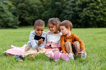 Naklejka na ściany i meble childhood, leisure and technology concept - happy children with smartphone sitting on blanket outdoors