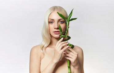 A blonde-haired blue-eyed young woman with smooth fresh skin holds a green bamboo branch near her face. Advertising of organic cosmetics. Skin and hair care products with natural composition.