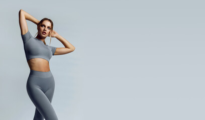Beautiful young woman with natural make-up and gathered hair, dressed in a gray sports uniform, posing in the studio on a gray background.Advertising sportswear and yoga wear. Healthy lifestyle, sport - Powered by Adobe
