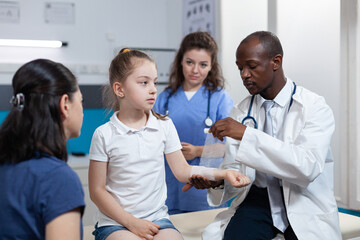 Obraz premium African american pediatrician doctor bandage broken bone of young child arm during medical consultation in hospital office. Girl patient with fractured hand having physiotherapy. Trauma recovery