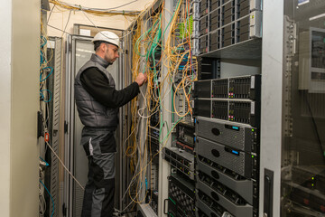 An engineer in a white helmet is measuring the level of an optical signal in a server room. A technician works near racks with network equipment and lots of wires.