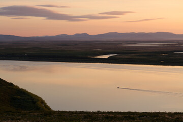 Obraz premium Evening view of the river valley. Summer arctic landscape. A boat is sailing along a wide river. In the distance tundra and mountains. The nature of Chukotka and Siberia. Anadyr River, Far East Russia