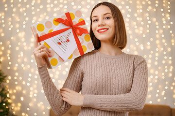 Portrait of young happy woman red lips holding wrapped gift box. Close up satisfied woman received present box. Festive Christmas lights background.