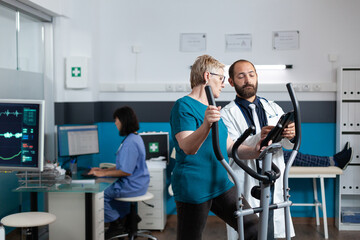 Senior patient using electrical bicycle for physical recovery while doctor holding digital tablet for healthcare. Woman with injury doing exercise activity for wellness and physiotherapy