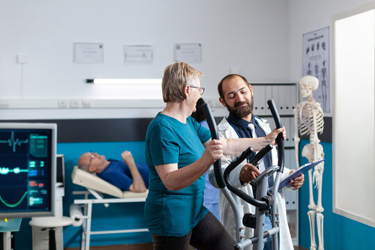 Doctor talking to old patient doing physical exercise to recover from muscle injury with bike. Aged woman using stationary bicycle for wellness and recovery at physiotherapy clinic.