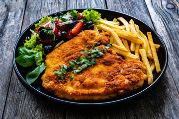 Breaded fried pork chop with French fries and fresh vegetables on wooden table
