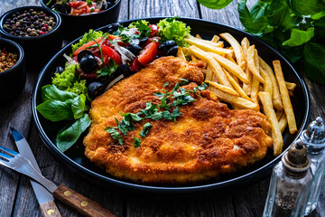 Breaded fried pork chop with French fries and fresh vegetables on wooden table
