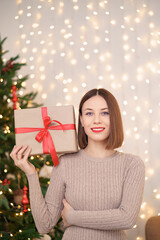 Portrait of young happy woman red lips looking at camera holding a wrapped gift box. Close up satisfied woman received present box. Festive Christmas lights background.