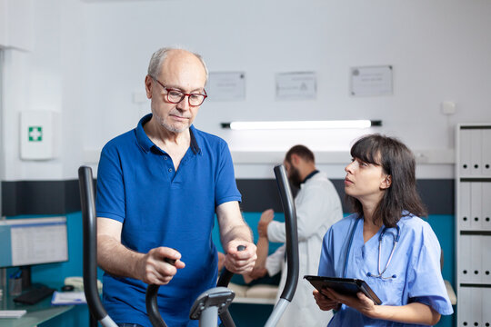 Aged man doing fitness exercise on stationary bicycle for recovery while nurse holding digital tablet. Patient with leg pain training for strength and healthcare. Adult at physiotherapy - Powered by Adobe