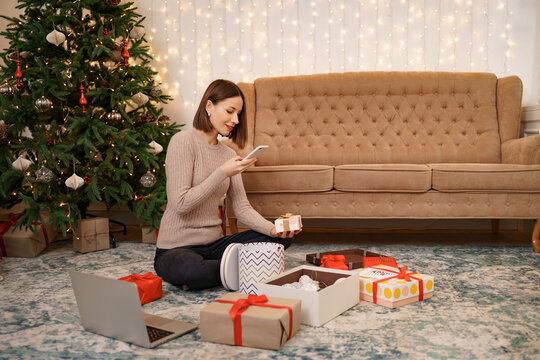 Woman wrapping Christmas gift and taking pictures on the phone while sitting in Christmas decorated living with a comfy sofa