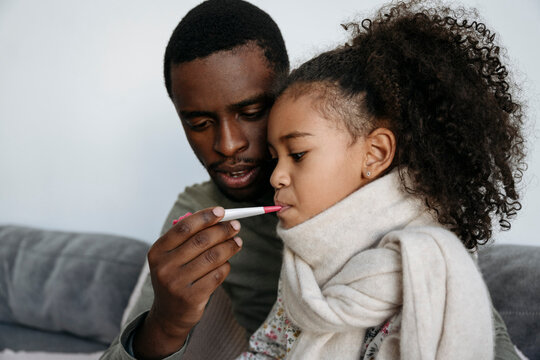 Young man measuring fever of daughter through thermometer in living room