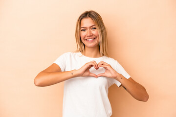 Young caucasian woman isolated on beige background smiling and showing a heart shape with hands.