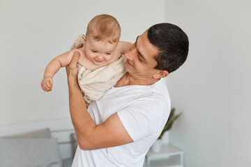 Satisfied attractive brunette man wearing white casual t shirt holding his infant daughter, posing at home in light room, spending time with his child with pleasure.